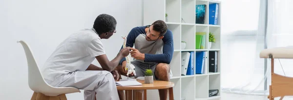 Réhabilitologue afro-américain mature et jeune patient pointant vers le modèle de la colonne vertébrale pendant la consultation, bannière — Photo de stock