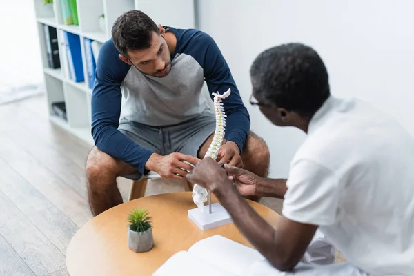 Médico afroamericano borroso hablando con paciente cerca del modelo de columna vertebral en el centro de rehabilitación - foto de stock