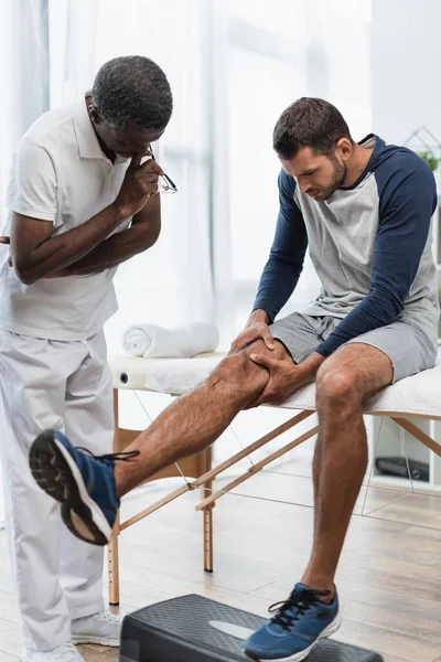 Junger Mann berührt Knie, während er auf Massagetisch in der Nähe eines afrikanischen amerikanischen Rehabilitologen sitzt — Stockfoto