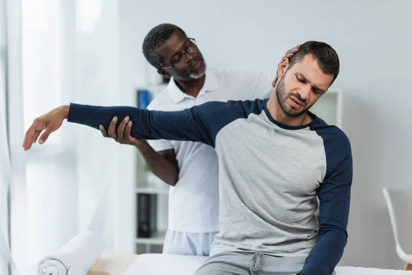 Mature african american rehabilitologist holding arm of young man frowning and suffering from pain — Stock Photo