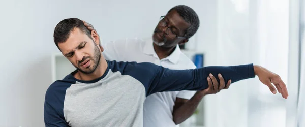 Joven frunciendo el ceño por el dolor mientras el rehabilitador afroamericano levanta su brazo, pancarta - foto de stock