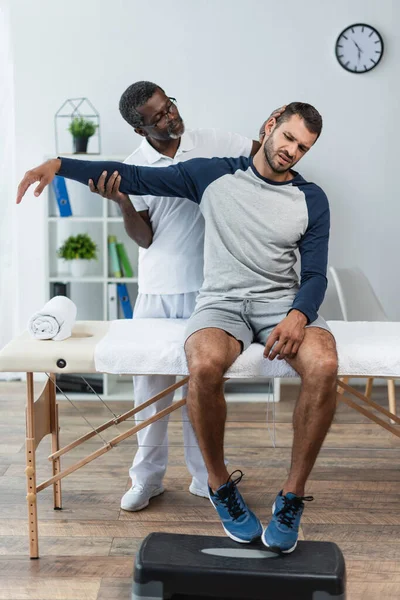 Young man frowning from pain while african american doctor examining him in rehabilitation center — Fotografia de Stock