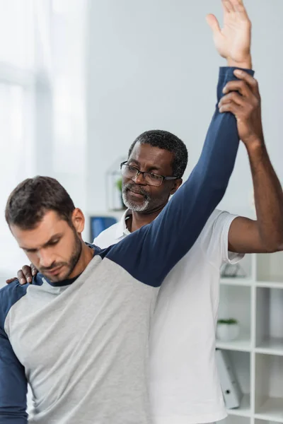 Mature afro-américaine médecin levant la main du jeune homme tout en l'examinant en centre de réadaptation — Photo de stock