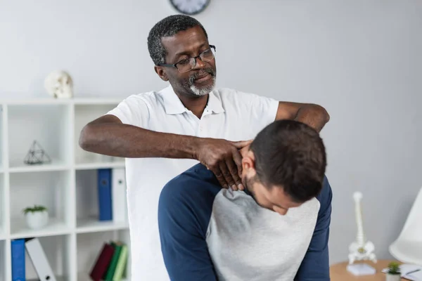 Mature african american physiotherapist examining neck of young man in rehabilitation center — Stock Photo