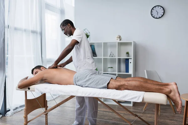 Молодой человек лежит на массажном столе во время массажа африканского американского реабилитолога — стоковое фото