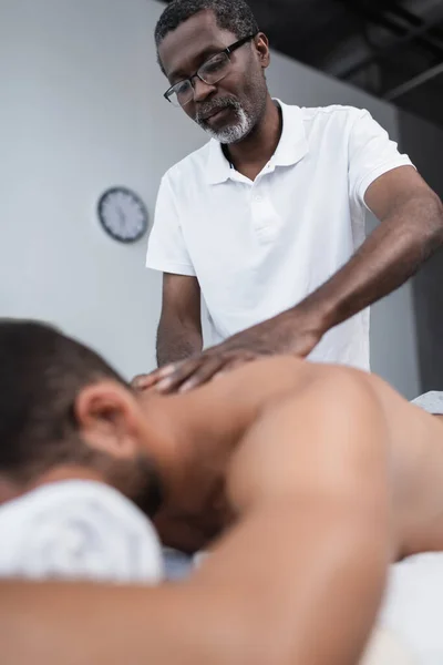 Vista de bajo ángulo del masajista afroamericano haciendo masaje de espalda durante la rehabilitación del hombre borroso - foto de stock