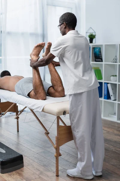 Riabilitologo afroamericano che allunga le gambe del giovane sul lettino da massaggio — Foto stock