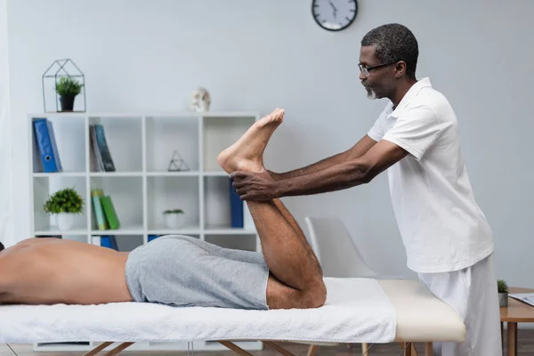Вид збоку афроамериканського фізіотерапевта, який лікує чоловіка, лежить на масажному столі — стокове фото