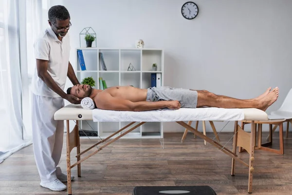 Vista completa del rehabilitólogo afroamericano tratando a joven en mesa de masaje - foto de stock