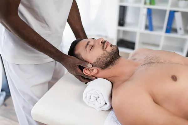 Jovem com olhos fechados deitado na mesa de massagem durante o tratamento de reabilitação pelo massagista afro-americano — Fotografia de Stock