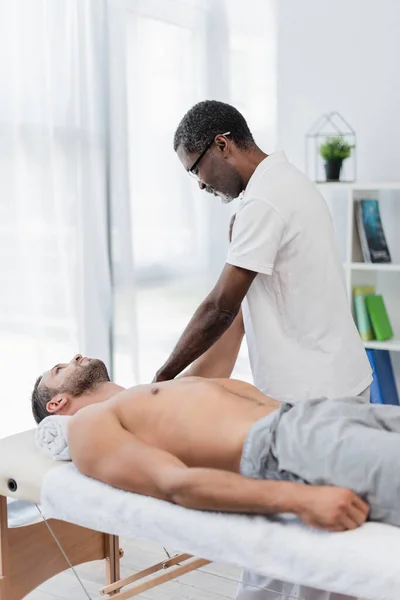 African american physical therapist massaging man in rehab center — Fotografia de Stock