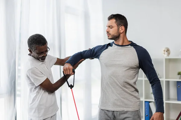 Mature african american trainer helping young man exercising with resistance band in rehabilitation center — Fotografia de Stock