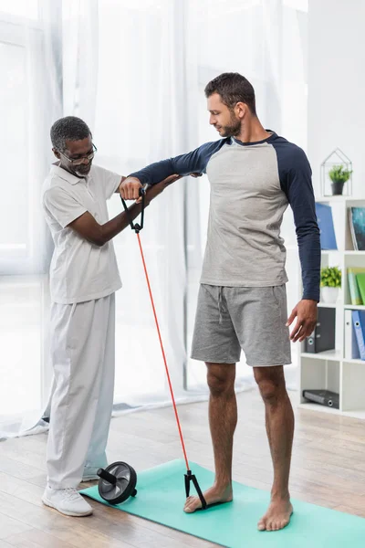 Mature african american physical therapist assisting barefoot man working out with resistance band — Stockfoto
