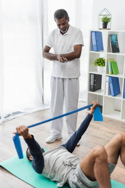 Rehabilitólogo afroamericano mirando a un joven entrenando con banda elástica mientras está acostado en una colchoneta de fitness - foto de stock