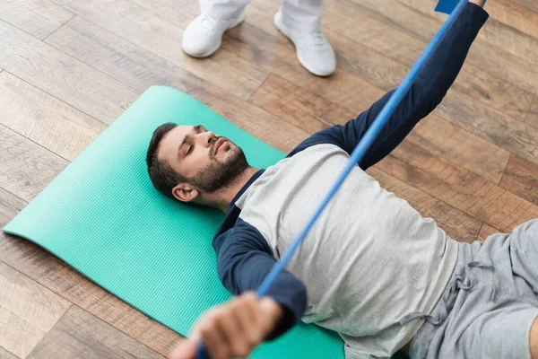 Jeune homme travaillant avec bande de caoutchouc tout en étant couché sur tapis de fitness près de réhabilitologue — Photo de stock