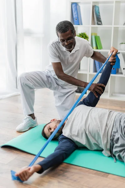 Rehabilitólogo afroamericano maduro ayudando a un joven a entrenar con una banda elástica en una esterilla de fitness - foto de stock