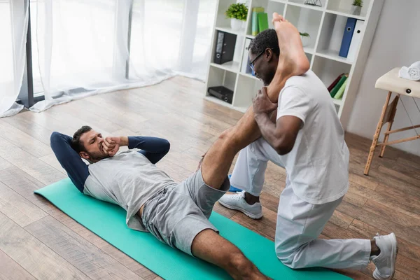 Young man feeling pain while african american rehabilitologist stretching his leg — Fotografia de Stock