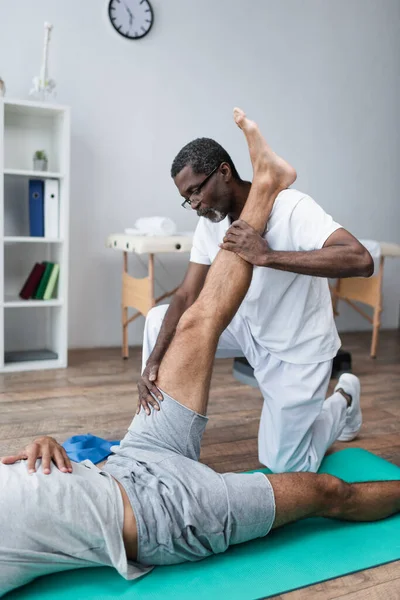 Riabilitologo africano allungamento gamba dell'uomo sdraiato sul tappeto fitness nel centro di riabilitazione — Foto stock