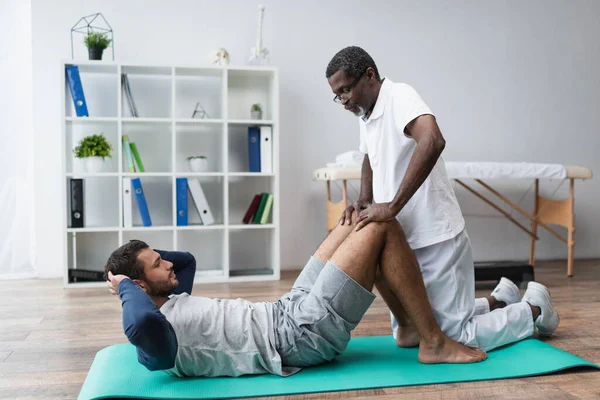 Afroamericano entrenador ayudar hombre haciendo abdominales ejercicio en centro de rehabilitación - foto de stock