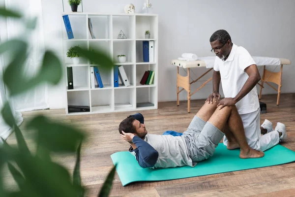 Entrenador afroamericano que ayuda al hombre a hacer ejercicio abdominales en centro de rehabilitación en primer plano borroso - foto de stock