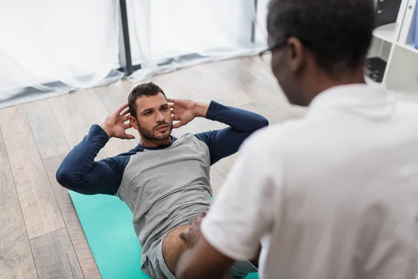 Desfocado afro-americano fisioterapeuta ajudando o homem fazendo abdominais exercício no tapete de fitness — Fotografia de Stock