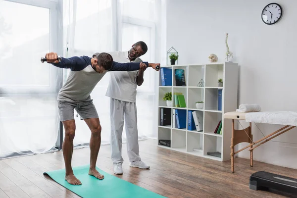 Зрілий афроамериканський реабілітолог допомагає молодому чоловікові тренуватися з гантелями на фітнес-маті — стокове фото