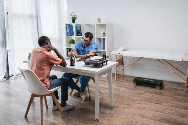 Physiothérapeute pointant avec un stylo tout en parlant à un homme afro-américain souffrant de douleurs au cou — Photo de stock