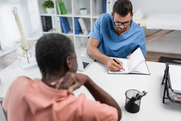 Joven rehabilitólogo escribiendo en cuaderno cerca borrosa afroamericano hombre que sufre de dolor de cuello - foto de stock