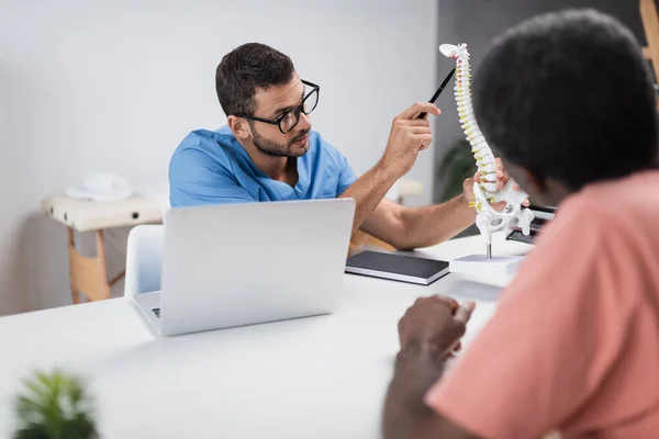 Physiotherapeut mit Brille zeigt auf Wirbelsäulenmodell nahe verschwommenem afrikanisch-amerikanischem Patienten — Stockfoto