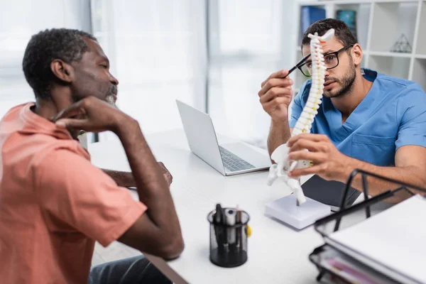 Rehabilitologist pointing at spine model near blurred african american patient suffering from neck pain — Stockfoto