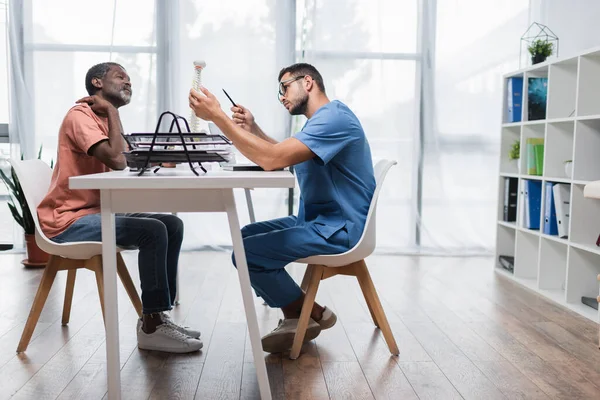 Seitenansicht des Rehabilitologen zeigt auf Wirbelsäulenmodell in der Nähe eines reifen afrikanisch-amerikanischen Mannes mit Nackenschmerzen — Stockfoto