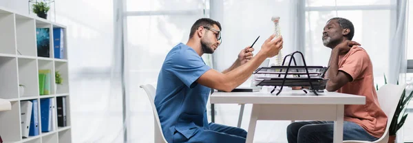 Médico en el centro de rehabilitación apuntando al modelo de columna vertebral cerca de paciente afroamericano que sufre de dolor de cuello, bandera - foto de stock