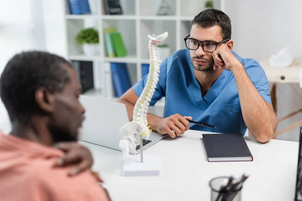 Joven rehabilitólogo en anteojos escuchando borroso afroamericano hombre en consultorio - foto de stock