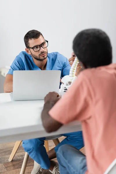 Rückansicht eines verschwommenen afrikanisch-amerikanischen Mannes in der Nähe eines Physiotherapeuten, der auf ein Wirbelsäulenmodell zeigt — Stockfoto