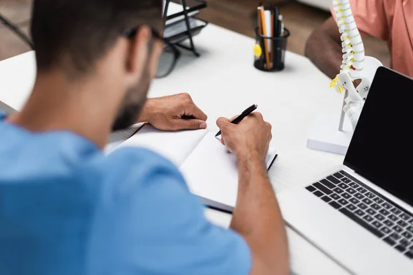 Medico offuscata scrittura in notebook vuoto vicino laptop e paziente afroamericano in centro di riabilitazione — Foto stock