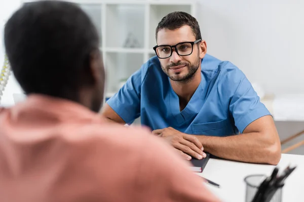 Lächelnder Reha-Arzt mit Brille hört verschwommenem afrikanisch-amerikanischem Patienten zu — Stockfoto