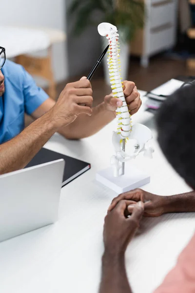 Vue partielle du médecin pointant vers le modèle de la colonne vertébrale près de l'homme afro-américain flou dans le centre de réadaptation — Photo de stock