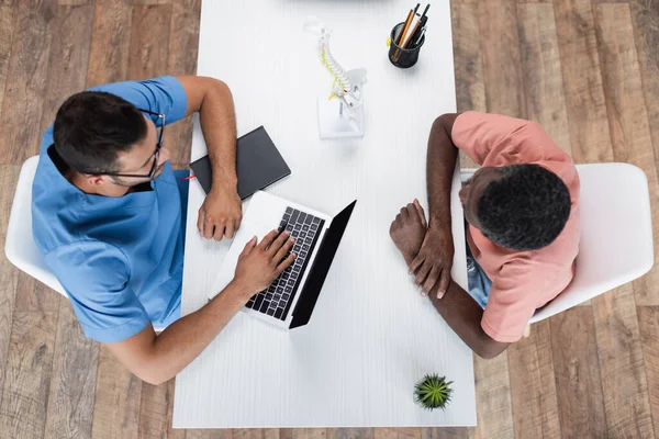 Top view of rehabilitologist and african american patient near laptop, notebook and spine model on desk — Stock Photo