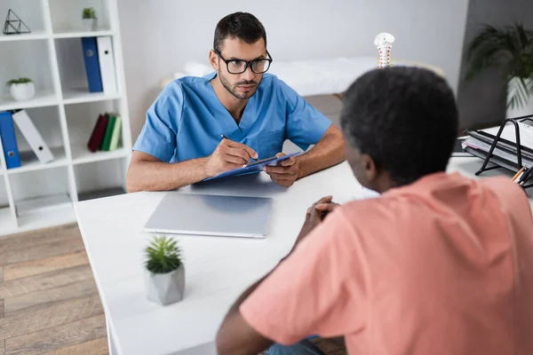 Jeune médecin écrivant sur presse-papiers tout en écoutant un patient afro-américain mature dans un centre de réadaptation — Photo de stock