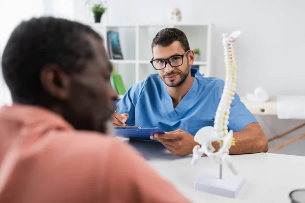 Positive rehabilitologist looking at camera while writing in notebook near blurred african american patient — Stock Photo