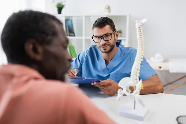 Positiver Rehabilitologe blickt in Kamera, während er in Notizbuch fast verschwommenen afrikanisch-amerikanischen Patienten schreibt — Stockfoto