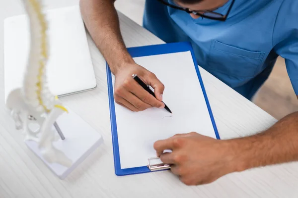 Ausgeschnittene Ansicht eines Rehabilitologen, der auf leeres Klemmbrett neben verschwommenem Wirbelsäulenmodell und Laptop auf Schreibtisch schreibt — Stockfoto