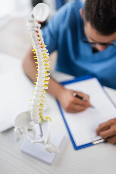 Selective focus of spine model near blurred rehabilitologist writing prescription on clipboard — Fotografia de Stock