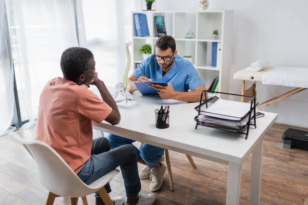 Joven rehabilitólogo escribiendo en portapapeles cerca de paciente afroamericano maduro - foto de stock