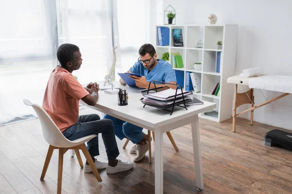 Mann mittleren Alters sitzt mit geballten Händen neben Reha-Arzt und schreibt Rezept auf Klemmbrett — Stockfoto