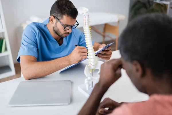 Jeune thérapeute de réadaptation écrit sur presse-papiers près flou afro-américain patient — Photo de stock