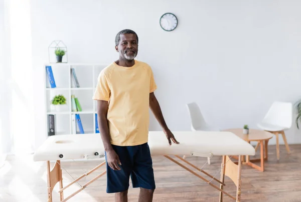 Positivo uomo afro-americano in piedi vicino al tavolo da massaggio nel centro di riabilitazione e guardando la fotocamera — Foto stock