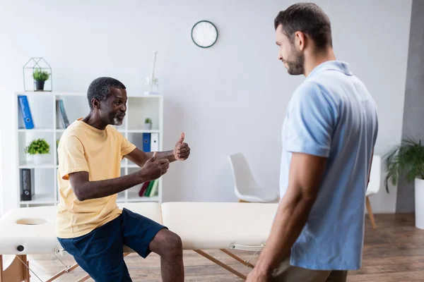 Mature african american man showing thumbs up while talking to rehabilitologist in clinic — Fotografia de Stock