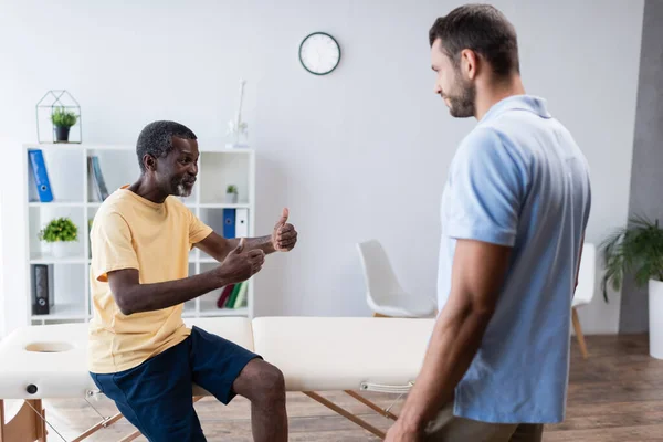 Homme mûr afro-américain montrant pouces levés tout en parlant à réhabilitologue à la clinique — Photo de stock