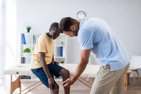 Young rehabilitologist pointing with hand at knee of african american patient — Stock Photo